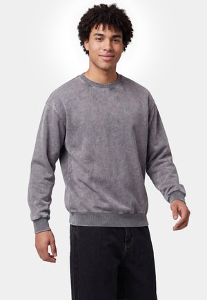 Junger Mann mit lockigem Haar, der einen grauen Langarm-Sweatshirt und schwarze Hose trägt, vor einem schlichten weißen Hintergrund.