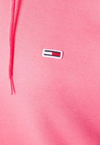 Růžová mikina s texturovanou látkou, na hrudi s malou vyšitou nášivkou v červené, bílé a modré. Nastavitelná kapuce na šňůrku.