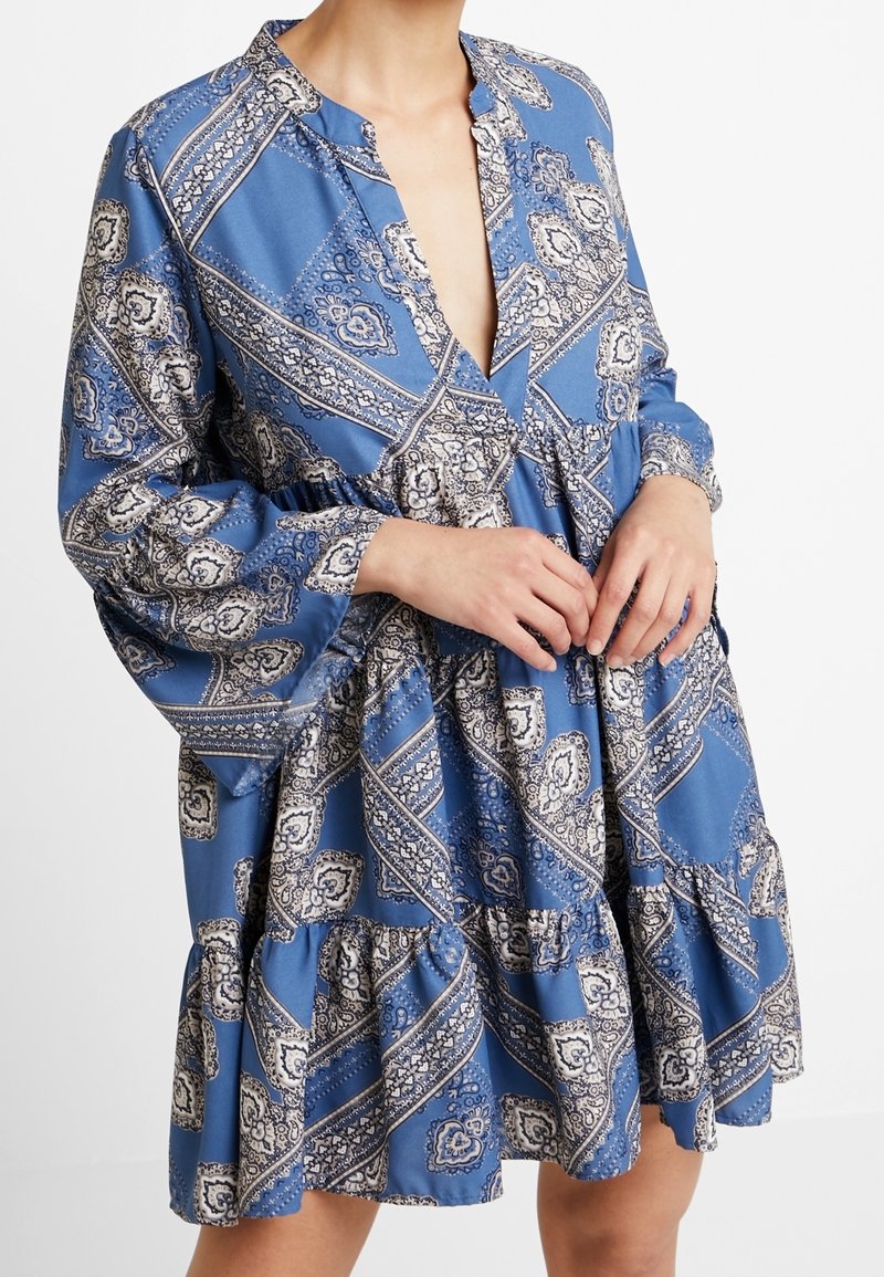 Femme portant une robe bleue avec des motifs floraux géométriques blancs et noirs et des manches longues bouffantes, les mains jointes à la taille.