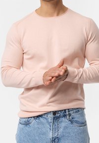 Dlouhý růžový svetr s kulatým výstřihem, žebrovanými manžetami a těsným střihem, v kombinaci s modrými denimovými džíny.