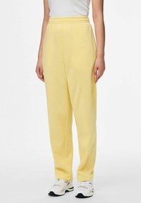 Vrouw in pastelgele sweatpants met elastische taille, witte sneakers en een witte mouwloze top, staand tegen een effen achtergrond.