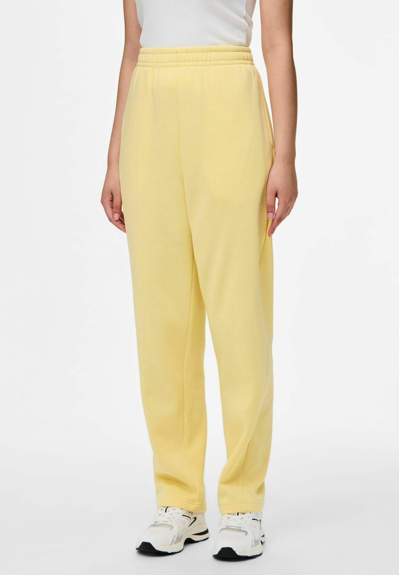 Vrouw in pastelgele sweatpants met elastische taille, witte sneakers en een witte mouwloze top, staand tegen een effen achtergrond.
