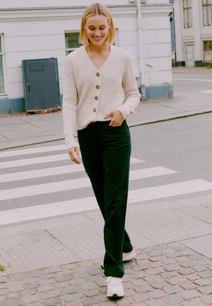 Krämfärgad ribbad cardigan med träknappar, parat med svarta raka byxor och vita sneakers, som går på ett övergångsställe.