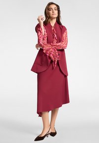 Gilet bordeaux sopra una blusa rosa a fantasia con scollatura arricciata. Presenta una gonna asimmetrica e scarpe con tacco basso color marrone.