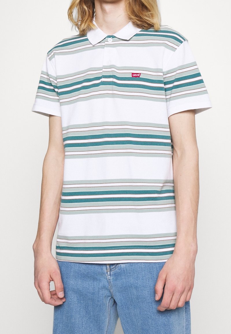 Personne portant un polo blanc à manches courtes avec des rayures horizontales vertes et grises et un jean bleu, debout devant un fond uni.