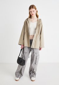 Béžová dažďová bunda s kapucňou, biely textúrovaný crop top, šedé cargo nohavice s vreckami a čierna taška so zlatými cvokmi; biele tenisky.