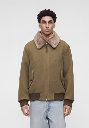 Jeune homme portant une veste vert olive avec un grand col en fourrure beige et un jean bleu clair, debout devant un fond clair uni.