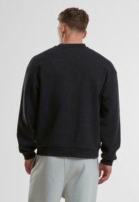 Homme de dos portant un sweat-shirt noir uni et un pantalon gris clair sur un fond neutre.