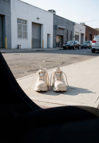 Weiße Sneakers mit offenen Schnürsenkeln, die auf einem Bürgersteig in der Nähe einer Straße mit Industriegebäuden und geparkten Autos liegen.