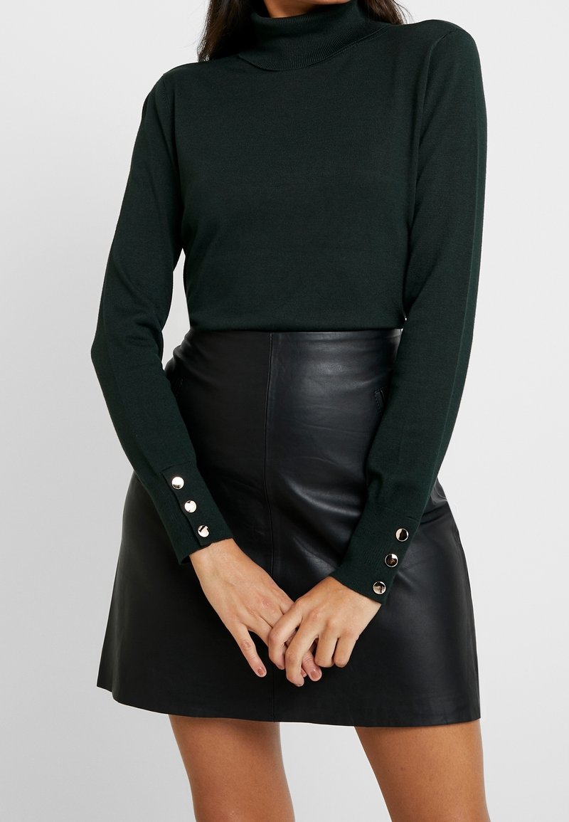 Grüner Strick-Turtleneck-Pullover mit langen Ärmeln und Knopfakzenten an den Bündchen, kombiniert mit einem hoch taillierten schwarzen Lederrock.