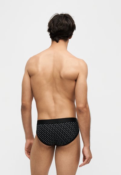 Homme vu de dos portant un slip noir avec un texte blanc en diagonale sur un fond gris clair uni.