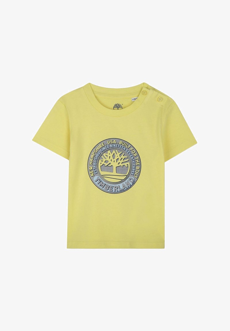 Camiseta de algodón amarilla con mangas cortas, que presenta un logotipo circular de Timberland en azul y gris y dos botones a presión en el hombro izquierdo.