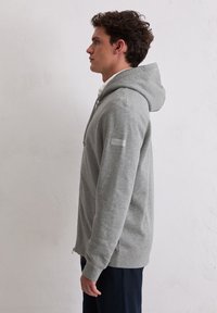 Felpa con cerniera grigia, dotata di tasca frontale, cappuccio con cordino e spacchi laterali. Presenta una patch in tessuto sulla manica sinistra. Texture liscia.