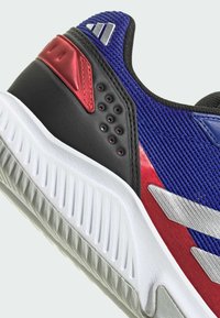 Chaussure de sport avec un dessus texturé bleu, un talon noir, des accents rouges et une semelle blanche avec des rainures pour l'adhérence. Décorée d'un logo argenté.