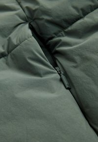 Jachetă îmblănită din material verde închis, având un fermoar vizibil și o textură subtilă, cu secțiuni cusute pentru izolație.