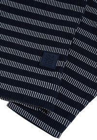 Pull à rayures bleu marine avec des lignes blanches alternées, une texture côtelée et une étiquette carrée bleu marine avec des accents de logo blancs en bas.