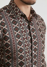 Chemise à boutons à motifs présentant des designs géométriques en rouge, vert, noir et crème. Fabriquée en tissu lisse avec un col.