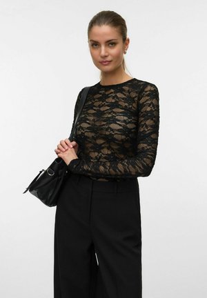 Femme aux cheveux tirés en arrière portant un haut noir en dentelle transparente à manches longues, un pantalon noir et un sac à épaule noir.