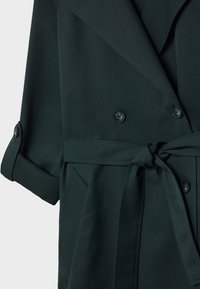 Ciemnozielony, dwurzędowy płaszcz z gładkiego materiału, z kołnierzem szalowym, podwiniętymi rękawami, paskiem oraz bocznymi kieszeniami.