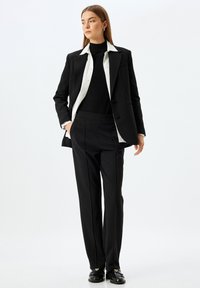 Veste noire avec col blanc sur un turtleneck noir, assortie à un pantalon noir ajusté. Mocassins noirs avec des embellissements. Texture lisse.