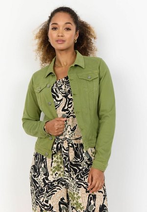 Woman with curly hair wears a green jacket over a patterned blouse and pants, posing confidently against a white background.