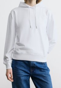 Personne portant un sweat à capuche blanc uni avec des cordons et un jean bleu, debout avec une main visible et l'autre derrière le dos.
