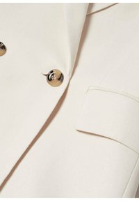 Blazer couleur crème avec un gros plan de deux boutons en écaille de tortue et un détail de manchette de manche pliée.