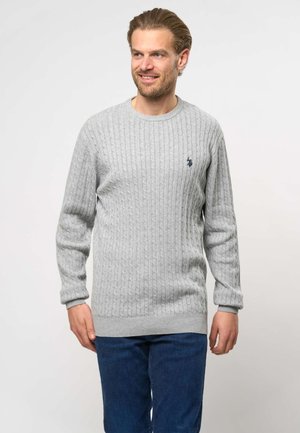 Homme aux cheveux châtain clair portant un pull en maille torsadée gris clair et un jean bleu foncé, souriant et regardant sur le côté.