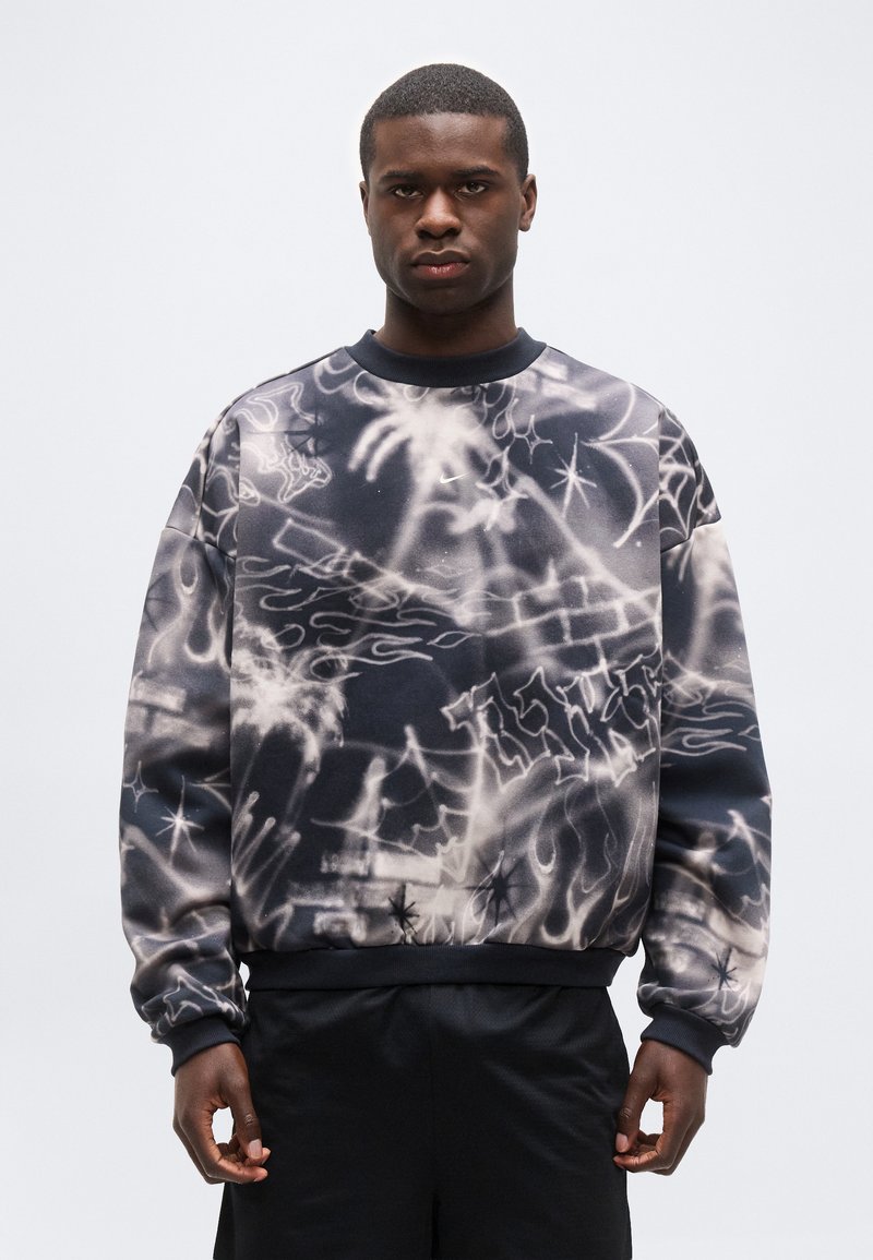 Άνδρας που φοράει φούτερ με μαύρα και άσπρα σχέδια, αφηρημένα και με φλόγες, στέκεται μπροστά σε απλό γκρι ανοιχτό φόντο.