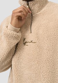 Pullover beige in pile con colletto alto e mezza cerniera. Presenta un tessuto morbido e texturizzato e un logo ricamato in verde.