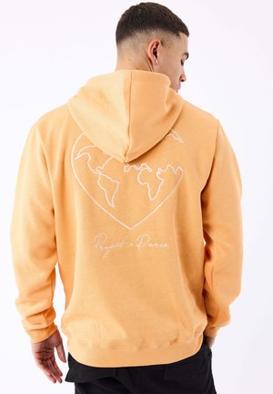 Uomo che indossa una felpa color pesca con cappuccio con un disegno bianco della mappa del mondo a forma di cuore e la scritta "Project x Paris" sul retro, visto di lato.