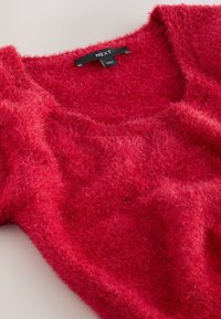 Crveni, pahuljasti džemper s mekanom teksturom i širokim izrezom, s oznakom na kojoj piše "NEXT." Tkanina ima lagano sjajni završetak.