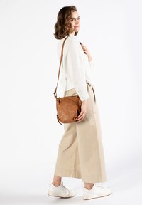 Bruine leren schoudertas met een zachte textuur, voorzien van een ritssluiting en een verstelbare schouderband. Draagt door een persoon in een witte trui en lichte broek.