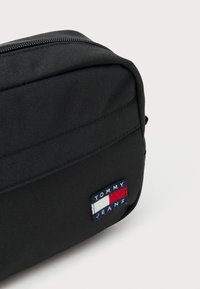 Fekete szövet tasak, texturált felülettel, cipzáras zárómechanizmussal és téglalap alakú kivitelben. Az oldalon egy színes "TOMMY JEANS" logó felvarró látható.