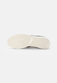 Black athletic shoe sole with zigzag tread pattern. Made of rubber, featuring a textured surface and a raised logo in the center.