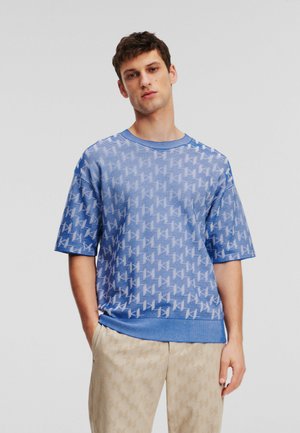 Camiseta de punto azul de manga corta con un patrón geométrico, rayas texturizadas y un cuello acanalado azul sólido. Combinada con pantalones de color beige claro.