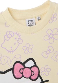 Felpa giallo chiaro con motivo floreale e fiocchi viola. Grafica di Hello Kitty in nero e rosa con dettaglio di fiocco rosa testurizzato.
