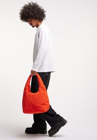 Borsa tote arancione imbottita con manico a taglio curvo, indossata da una persona con una camicia bianca a maniche lunghe e pantaloni neri, su uno sfondo semplice.