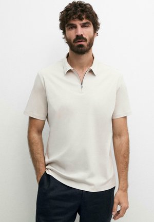 Mand med krøllet hår og skæg iført en beige poloshirt med kort ærme og lynlås og mørke bukser, stående med den ene hånd i lommen.