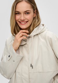 Lichtgewicht beige jack met een capuchon, voorzien van een ritssluiting, verstelbare trekkoorden en knoopmanchetten. Glad van textuur met een subtiele glans.