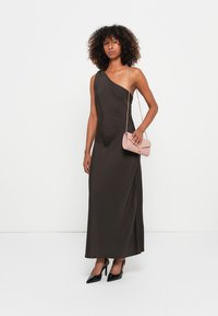 Robe longue marron à une épaule avec une texture lisse et élégante ; le mannequin tient une pochette rose avec une fine bandoulière, accompagnée de talons noirs.