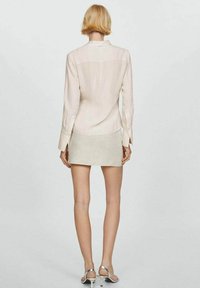 Donna con capelli biondi corti, che indossa una blusa rosa chiaro a maniche lunghe, una minigonna beige e scarpe con tacco alto argento, in piedi contro uno sfondo semplice.