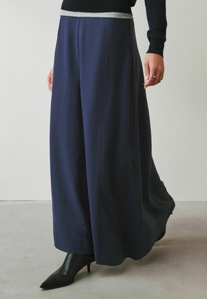 Person mit weiten marineblauen Hosen, schwarzen Stiefeletten mit Absatz, schwarzem Langarmoberteil und silbernem Ring an der rechten Hand, stehend in Innenräumen.