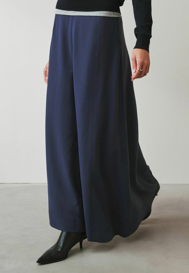 Personne portant un pantalon large bleu marine, des bottines noires à talons, un haut noir à manches longues, et une bague argentée à la main droite, debout à l'intérieur.