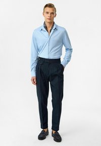 Lichtblauw overhemd met lange mouwen en een button-down kraag, gecombineerd met donkere geplooide broeken. De broeken hebben een unieke gespaccent.