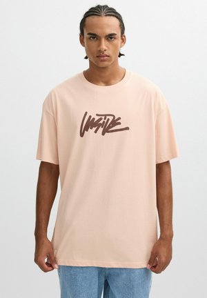 Camiseta de gran tamaño en rosa claro, hecha de un tejido suave, con un logotipo gráfico estilizado en marrón oscuro en la parte delantera. Mangas cortas y escote redondo.