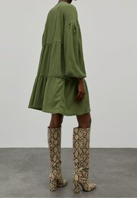 Vihreä teksturoitu mekko, jossa on ilmapallo-hihat ja kerroksittainen muotoilu, yhdistettynä polven ylle ulottuviin, paksupohjaisiin saappaisiin, joissa on käärmeennahkakuosi.