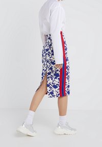 Midi sukně s květinovým vzorem v bílé, modré a červené barvě, se bočním rozparkem a kontrastním červeným pruhem. V kombinaci s kovovými stříbrnými teniskami.