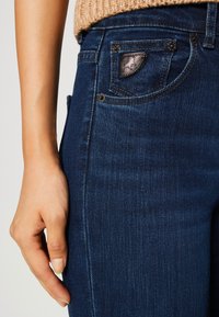 Tmavě modré džíny z denimu s přiléhavým designem, které mají malou náprsní kapsu s vyraženým detailem a odkrytou nití podél švů.