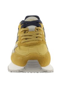 Sneaker giallo con tomaia in camoscio e tessuto, lacci blu navy, suola intermedia bianca e suola in gomma grigia. Presenta dettagli testurizzati e un patch con logo.